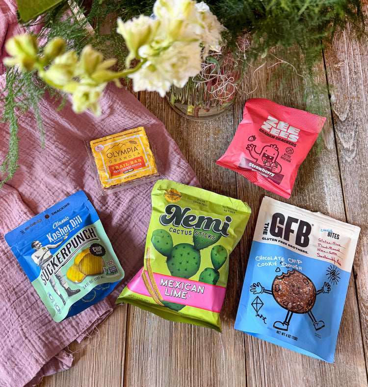 Five snack packets on a wooden surface with flowers and a pink cloth.