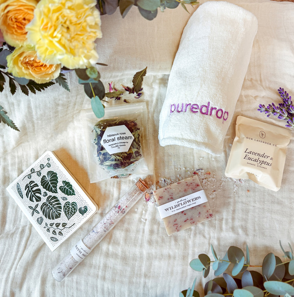 Collection of bath and body products including a towel, soap, and floral steam on a textured surface with flowers.